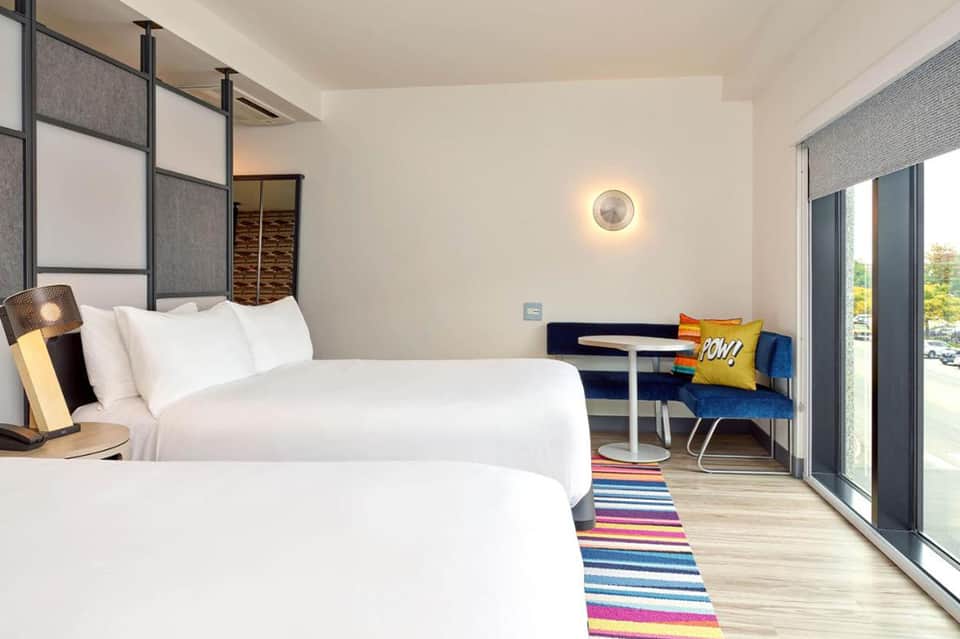 Modern hotel bedroom with two queen beds, colorful striped rug, blue seating area, and large windows