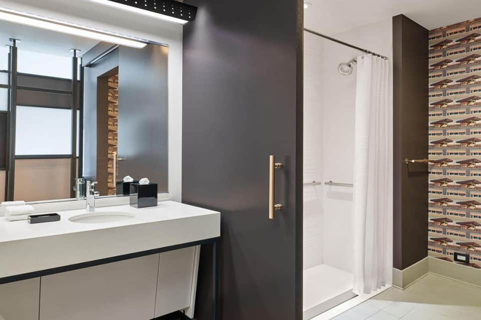 Modern hotel bathroom with white sink, dark wall panels, glass shower enclosure, and decorative tile accent