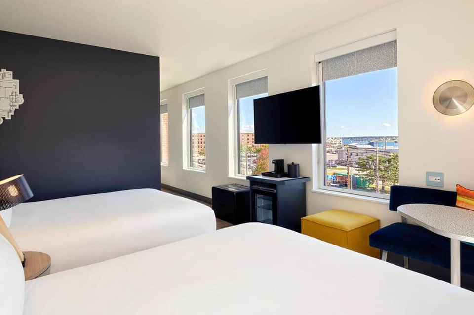 Modern hotel room with two beds, dark accent wall, flat-screen TV, and ocean city view windows