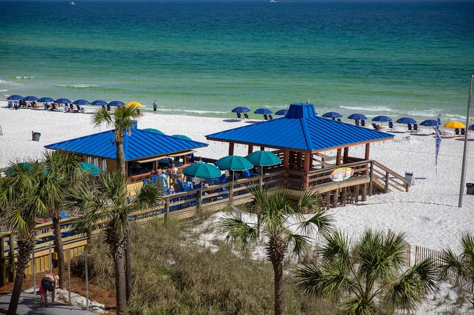 Beach pavilions with blue roofs, umbrellas, and palm trees along sandy shoreline