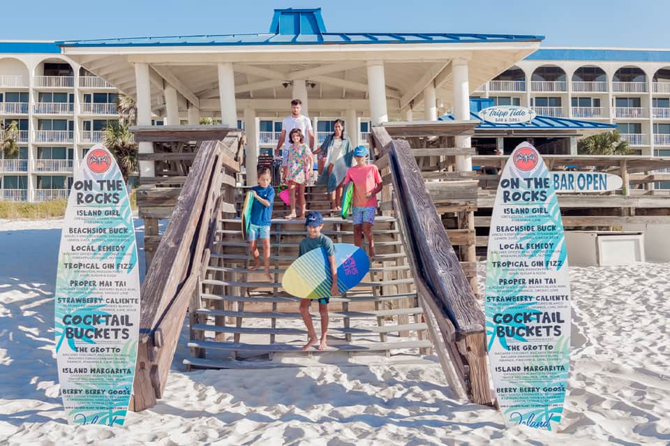 Family emerging from beach pier with colorful surfboard and beachfront resort in background