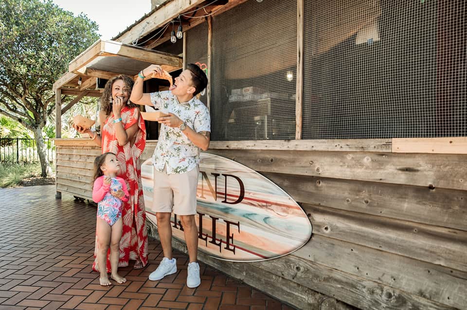 Family with surfboard outside beachside cabana with mesh screening and wooden railings