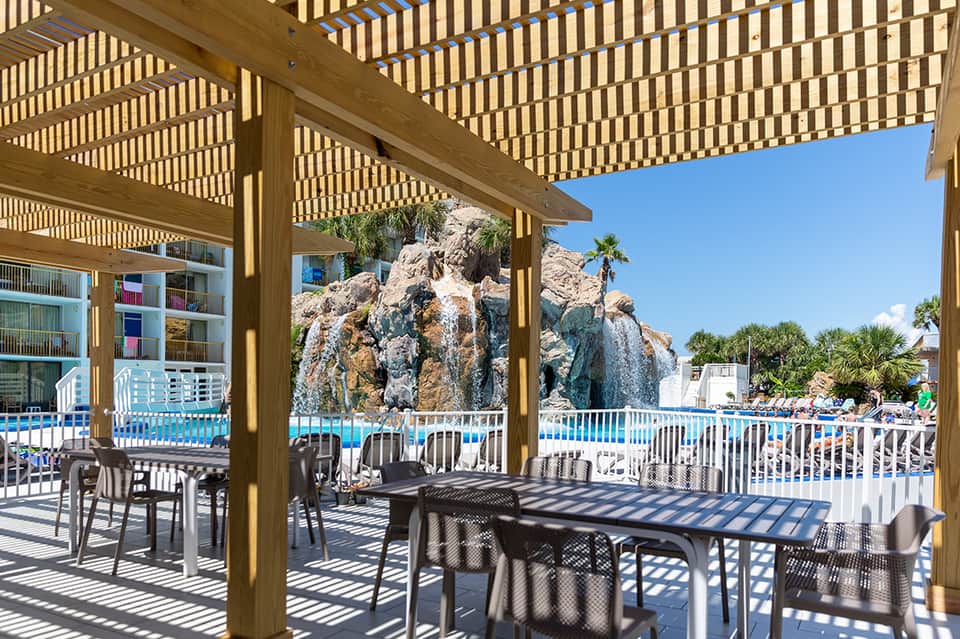 Shaded resort patio with wooden pergola, dining tables, and rocky landscape feature