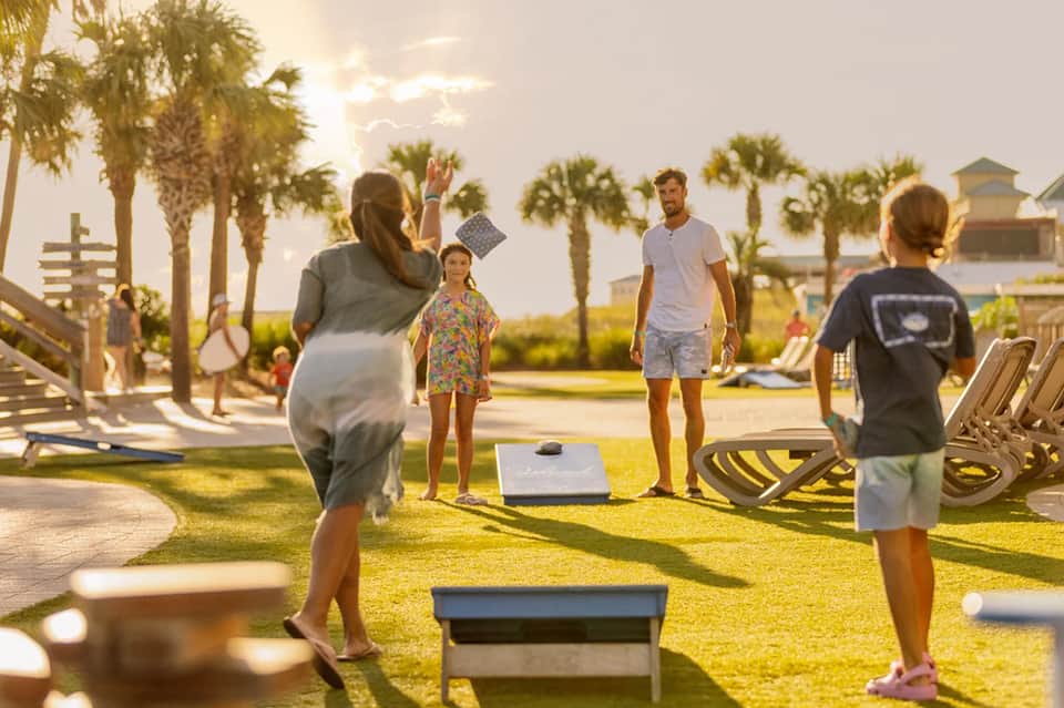 Family playing cornhole on resort lawn with palm trees and ocean view at sunset