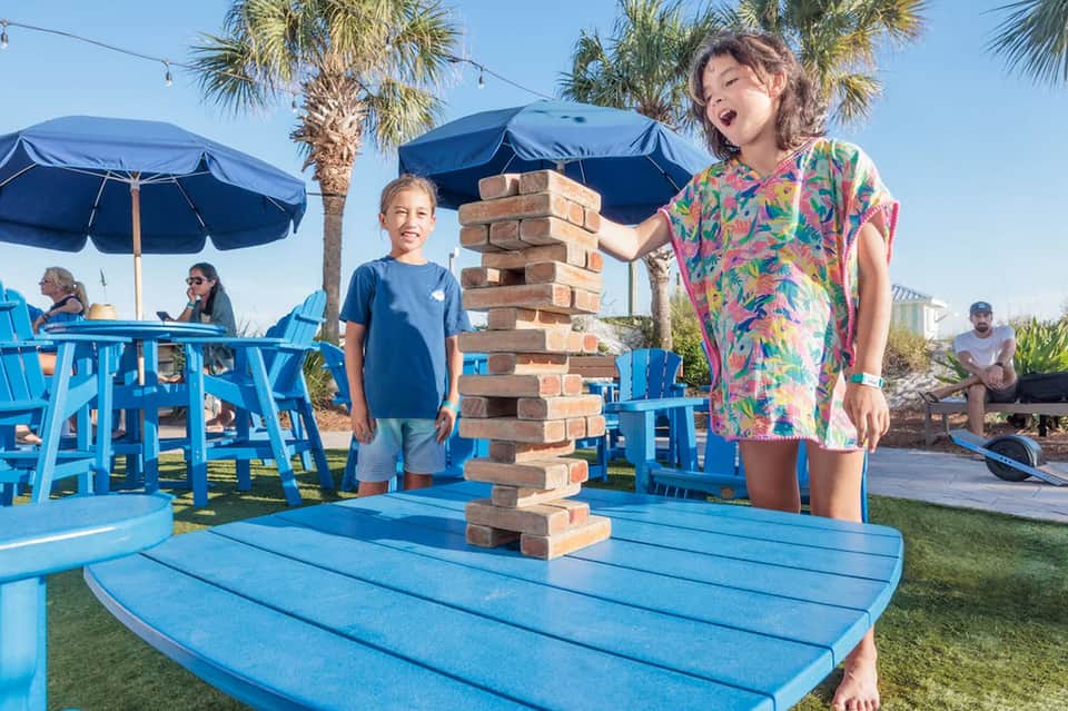 Family enjoying games by pool area with blue furniture, umbrellas and palm trees on sunny resort lawn