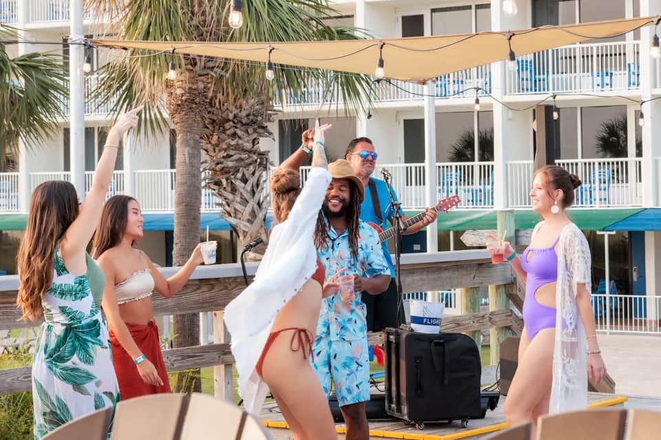 Beach musicians entertaining guests under shaded area with palm trees and resort building