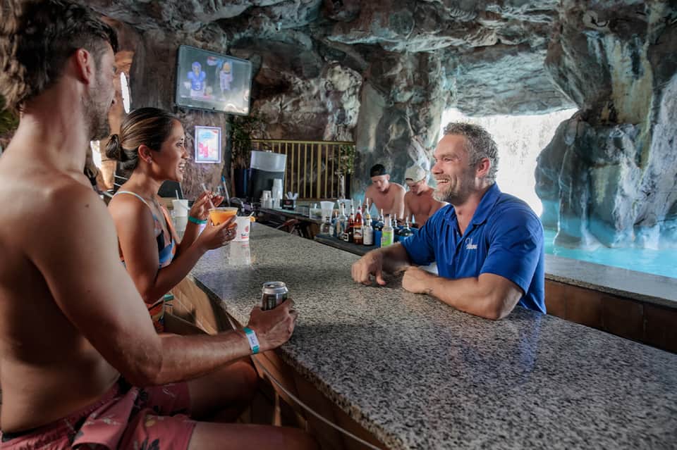 Beach bar built into cave with customers enjoying drinks, water view backdrop, and casual resort setting