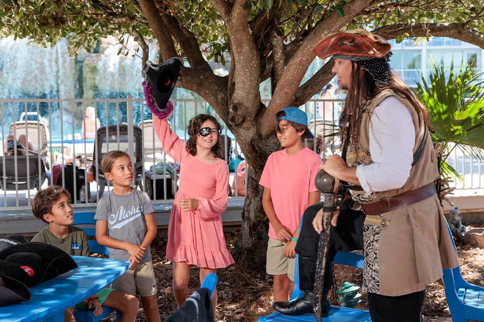 Family enjoying pirate character entertainment under large tree by resort pool area