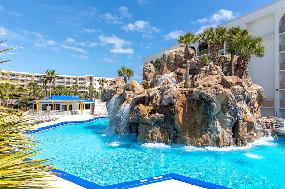 Resort pool with rocky waterfall feature, blue water, palm trees, and multi-story accommodations