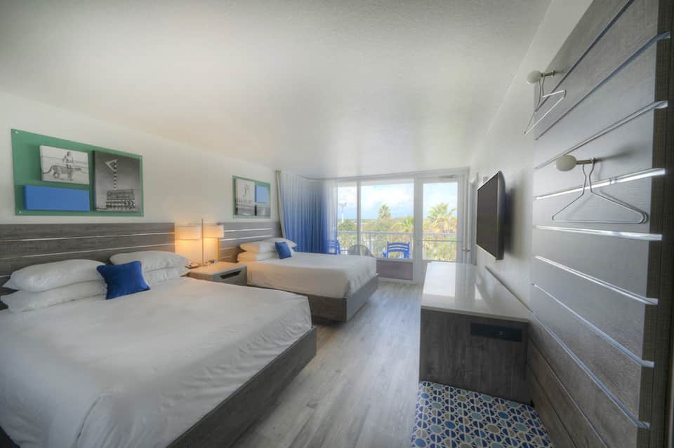 Modern hotel room with twin beds, ocean view balcony, wall-mounted TV, and blue accent pillows