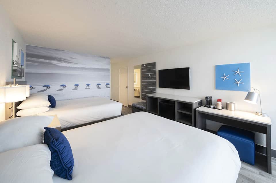 Modern hotel room with two queen beds, beach-themed wall mural, flat-screen TV, and coastal décor