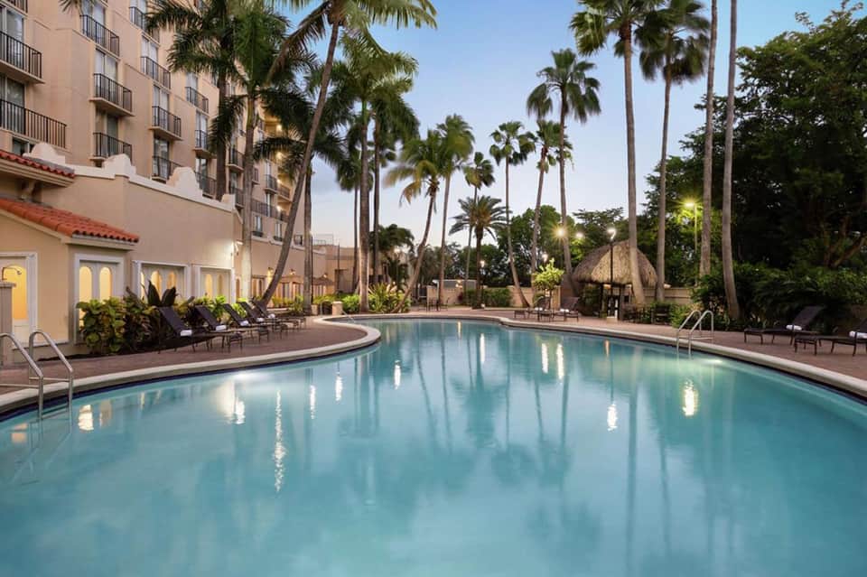 Resort pool at dusk with manicured landscape, palm trees, and adjacent hotel building