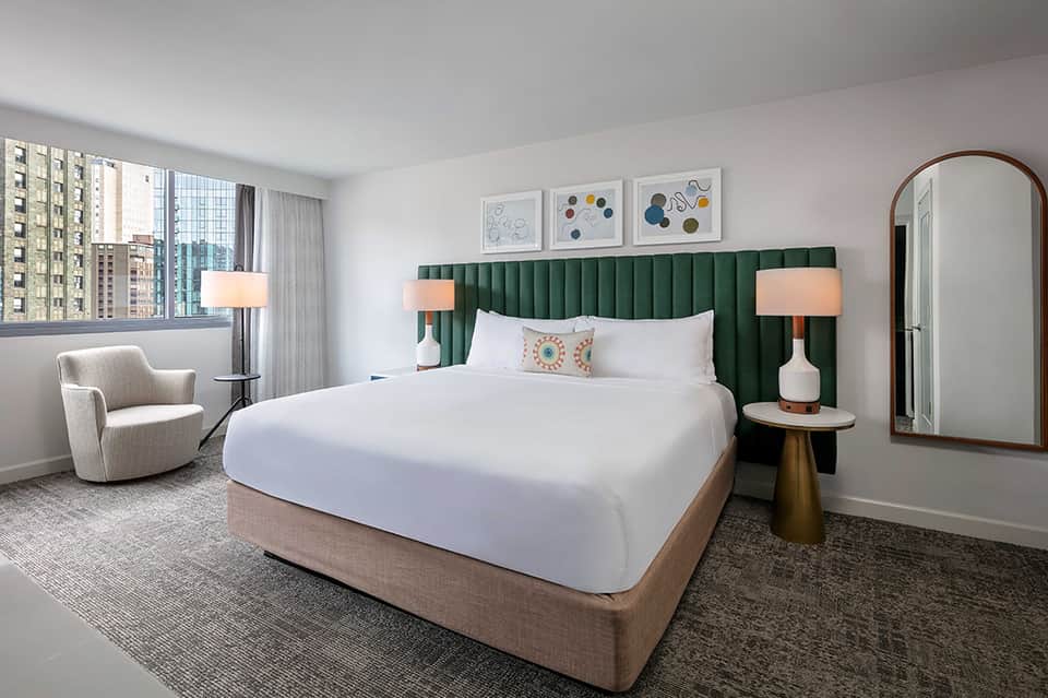 Modern hotel bedroom with green velvet headboard, white bedding, wood furniture, and city views