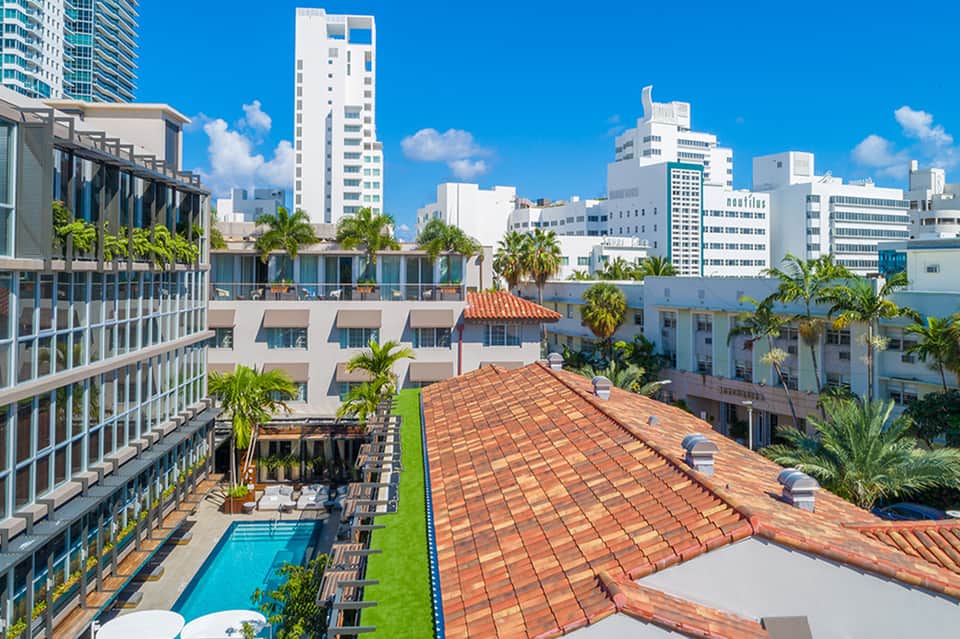 Urban rooftop pool area with terracotta roof, palm trees, modern high-rise hotels, and clear blue sky