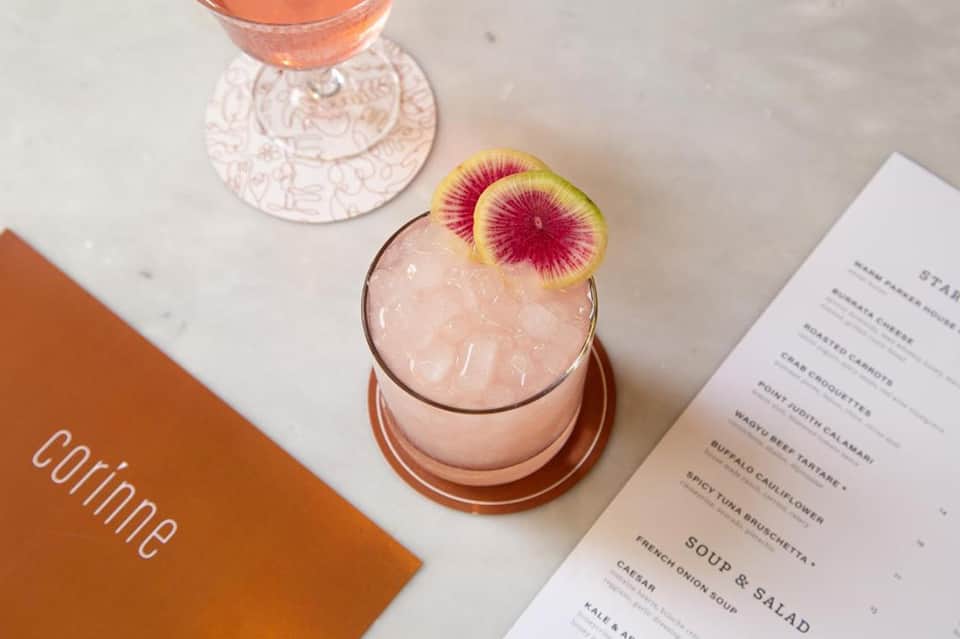 Elegant pink cocktail with watermelon slice garnish next to menu and place setting on white marble table