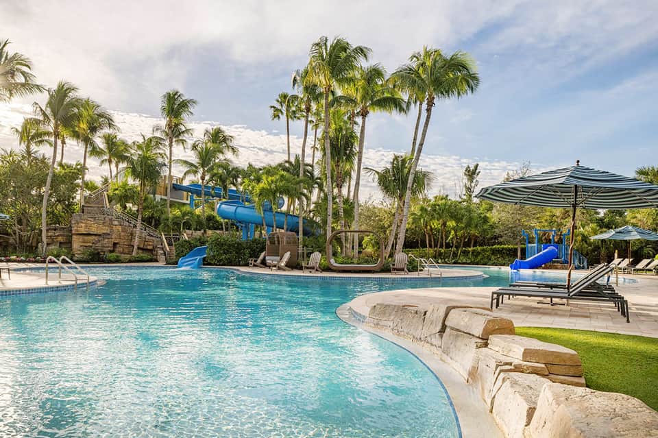 Tropical resort pool with blue water slides, palm trees, and lounge chairs with umbrellas