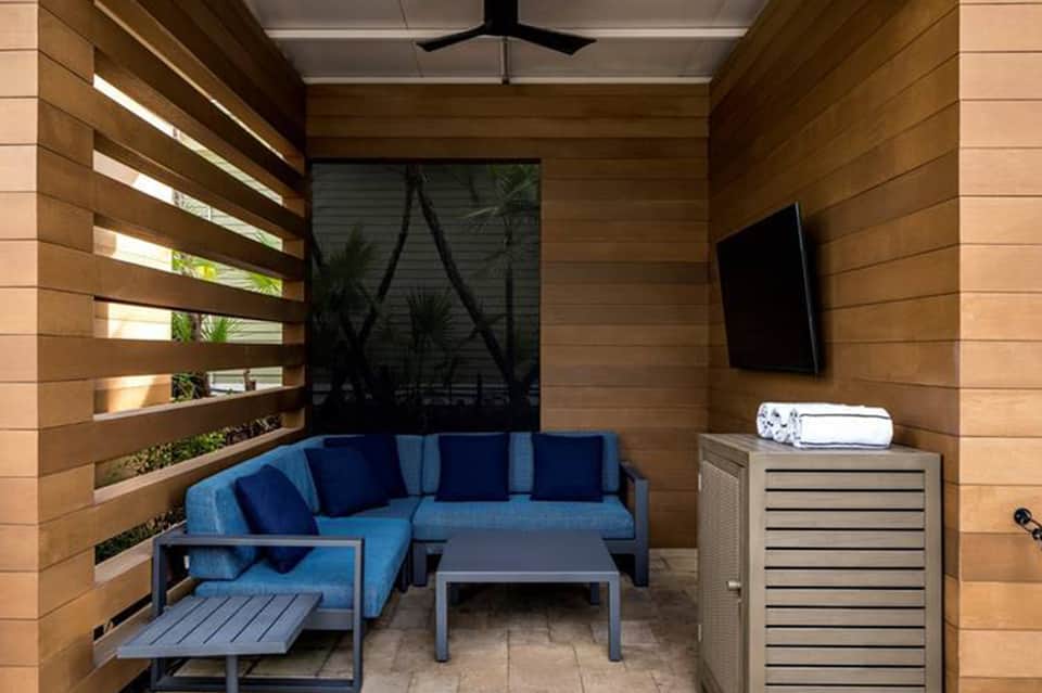 Private patio cabana with blue sectional sofa, wood walls, window slats, and TV mounted on wall