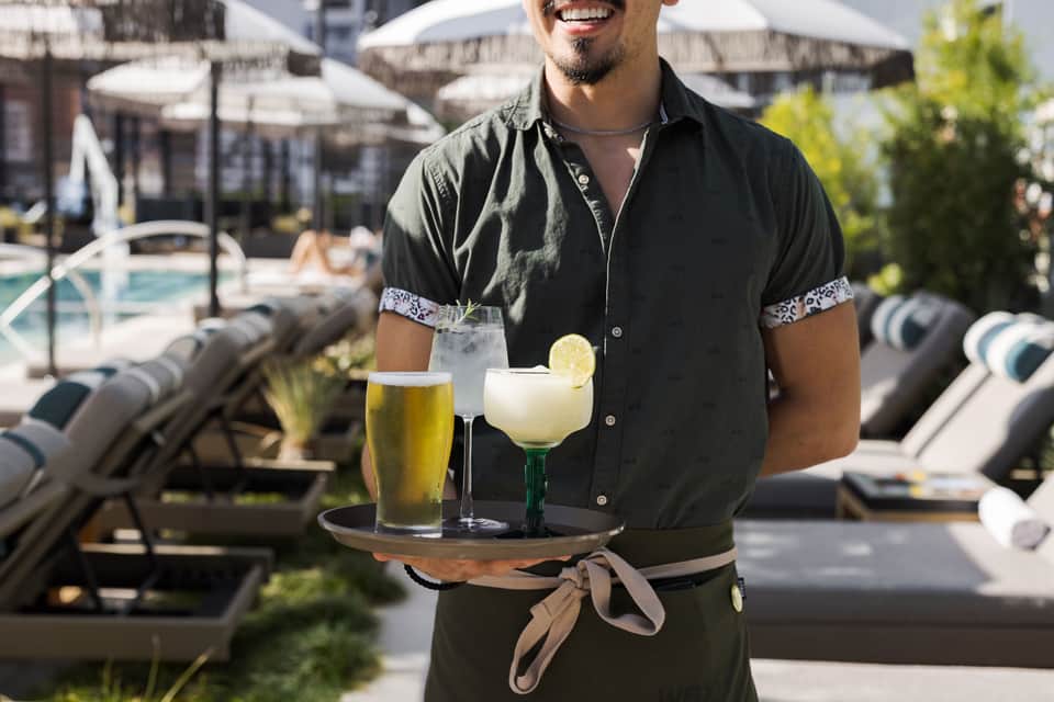 Smiling server holding drinks poolside with loungers and white umbrellas in background