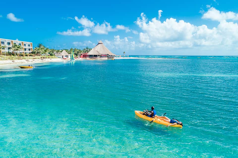 Kayaker in turquoise ocean with beach resort, tiki hut, and palm trees in background