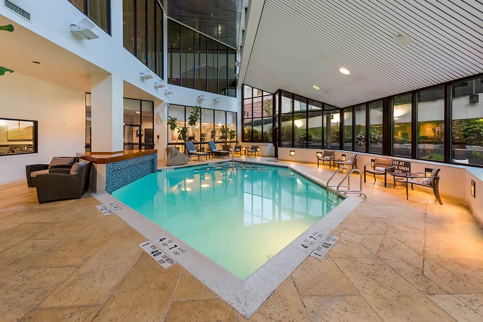 Modern indoor heated pool with turquoise water, floor-to-ceiling windows, lounge seating, and plants