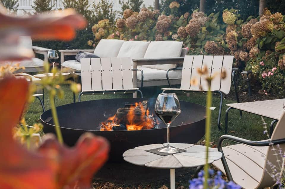 Fire pit seating area with white Adirondack chairs, wine glass, and garden backdrop