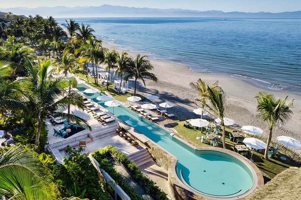 Beachfront resort with curved pool, loungers, palms, and distant mountain view