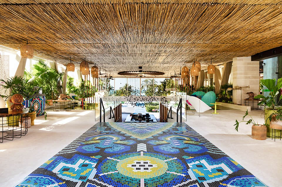 Grand resort lobby with decorative blue mosaic floor, woven ceiling, tropical plants, and water features