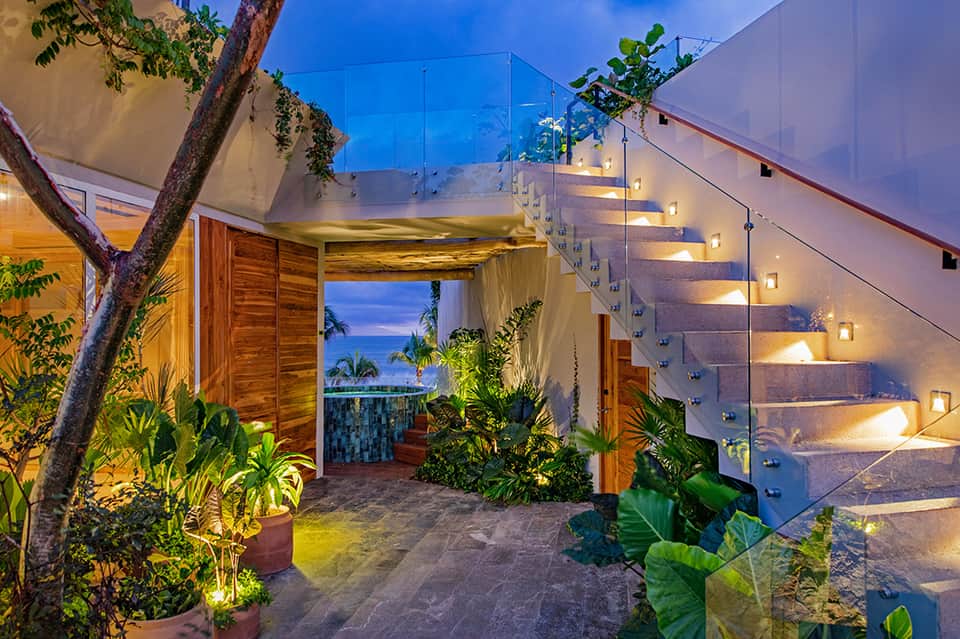 Modern villa staircase with ocean views, glass railings, tropical plants, and evening lighting