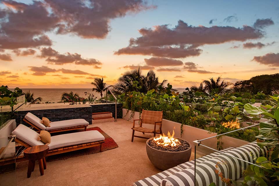 Beachfront fire pit lounge at sunset with loungers, chairs, palms, and ocean horizon