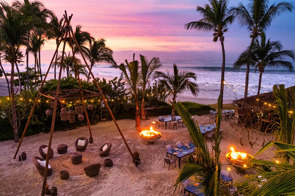 Beach bonfire at sunset with palm trees, lounge seating, and pink-purple sky over ocean