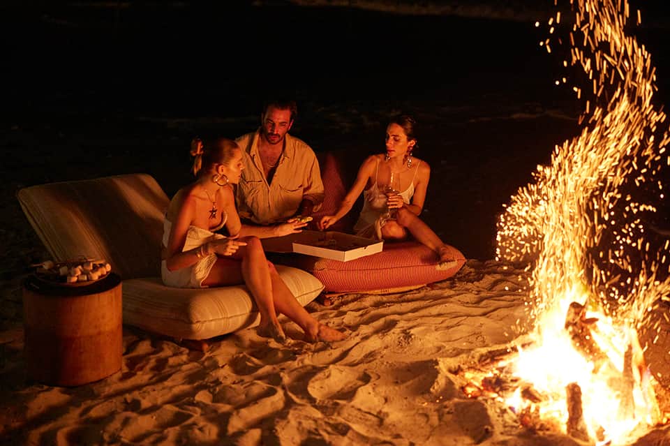 Group gathered around bonfire on beach at night with drinks and casual seating