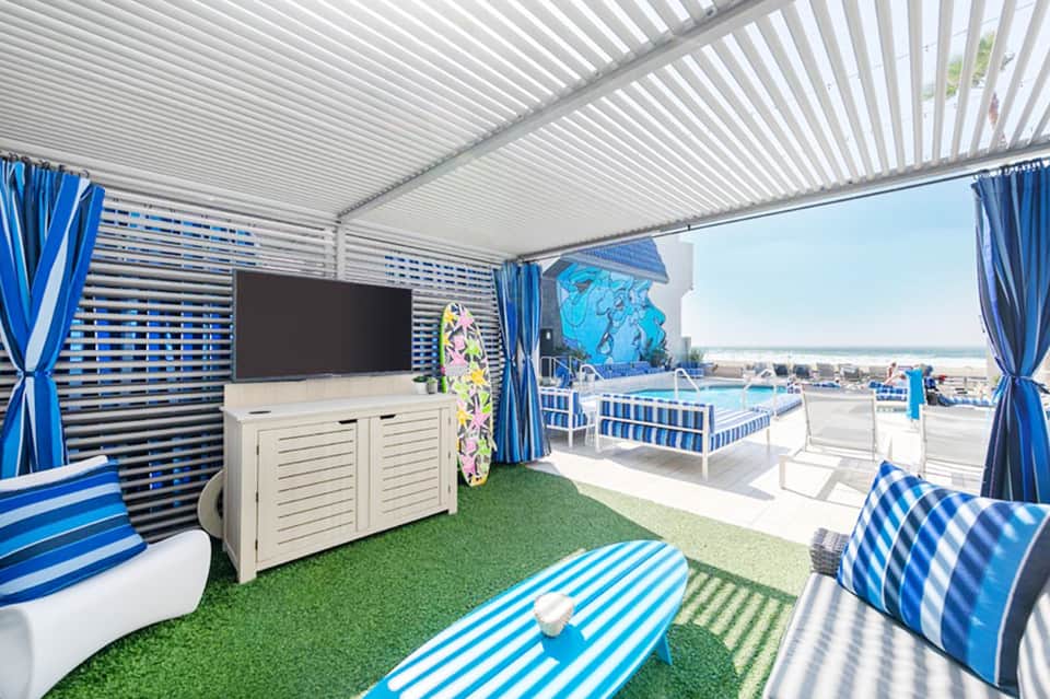 Covered beach cabana with blue striped furnishings, TV, and ocean view beyond railing