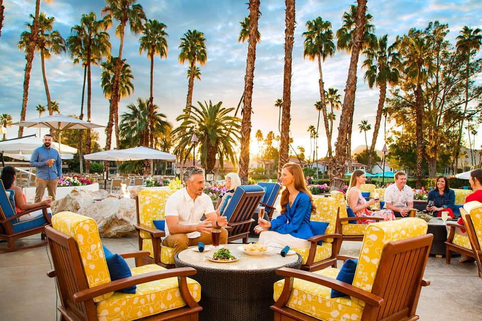 Outdoor seating area with guests in yellow and blue chairs, palm trees, and mountains