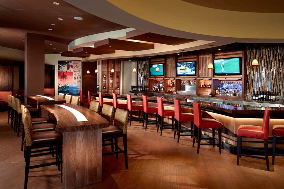 Upscale resort bar lounge with dark wood, red chairs, multiple TV screens, and curved ceiling