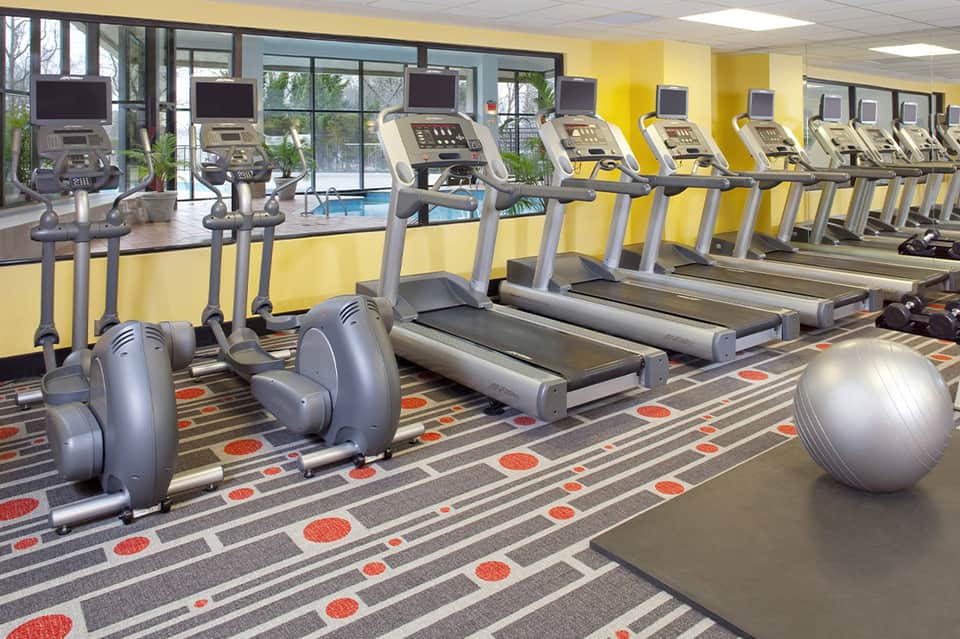 Bright fitness center with yellow walls, treadmills, elliptical machines, and pool view