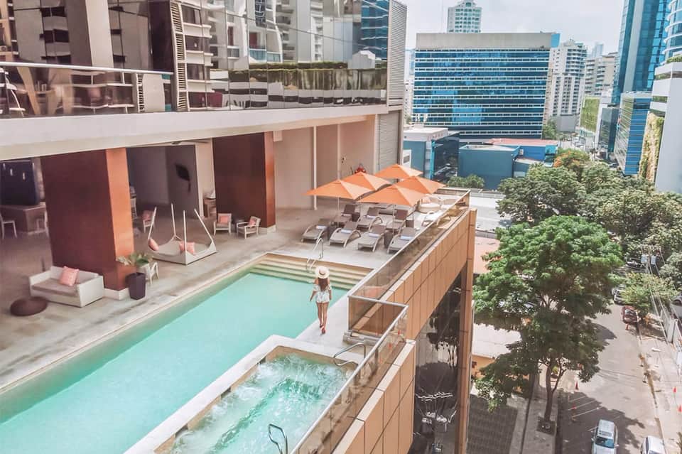 Elevated rooftop pool deck with orange umbrellas, lounge chairs, hot tub, and downtown city skyline views
