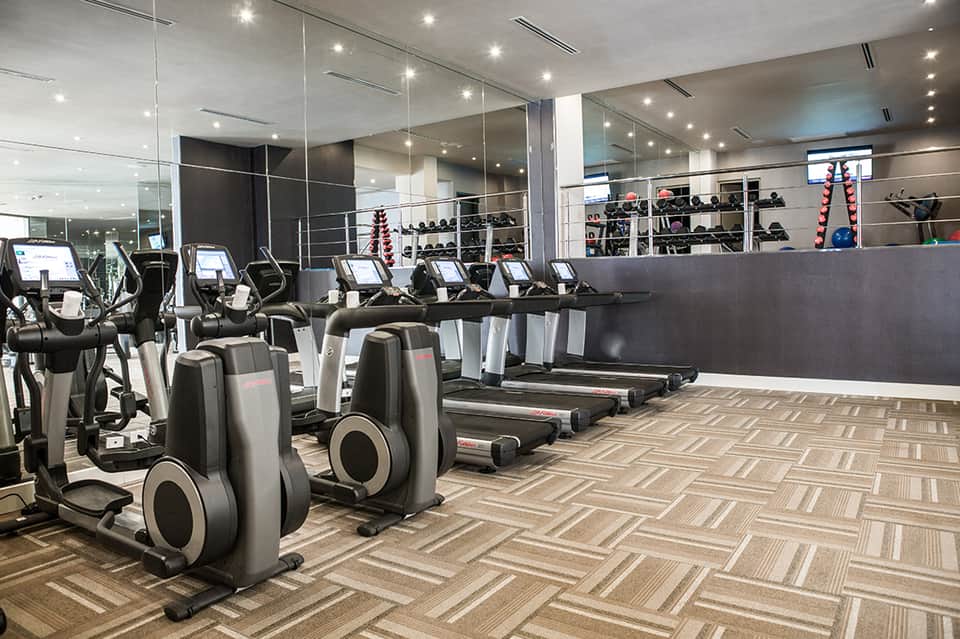 Modern gym with rows of cardio equipment, mirrors, weights, and contemporary flooring