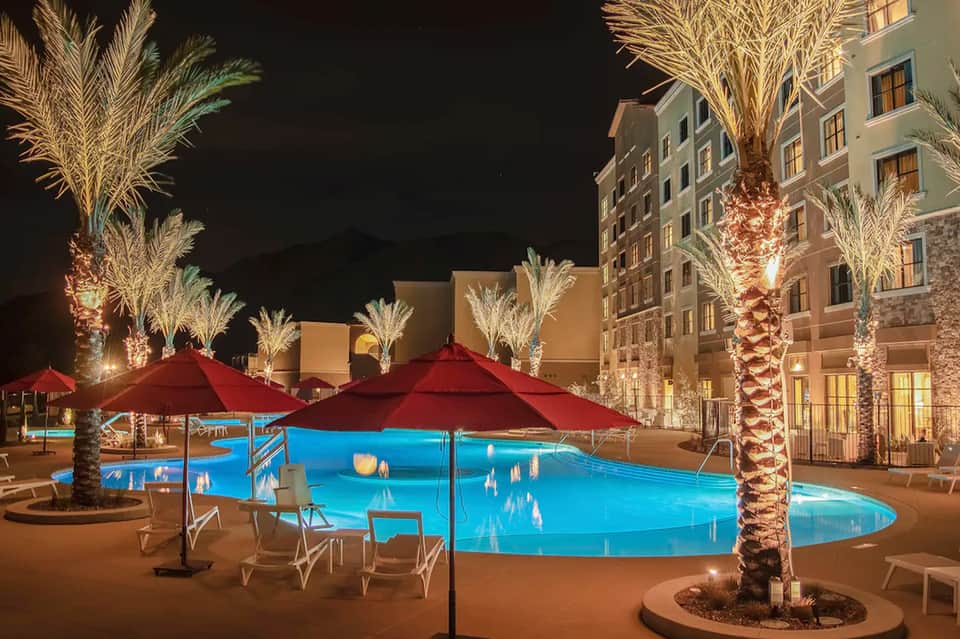 Lit resort pool at night with red umbrellas, palm trees, and multi-story hotel building