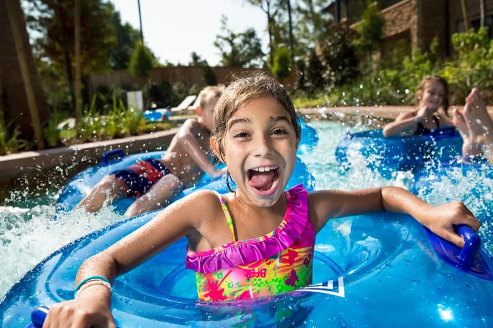 Happy children playing in bright blue swimming pool with water splashing and families in background