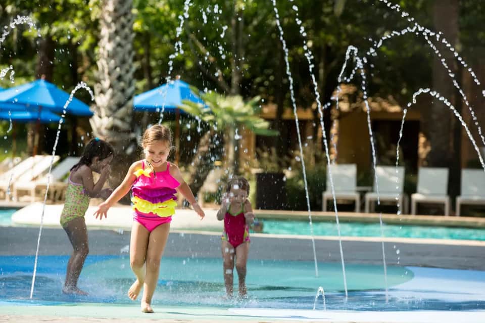 Children playing in splash pad fountains near pool with blue umbrellas, lounge chairs, and lush trees in background.