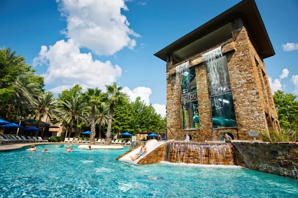 Resort pool with turquoise water, waterfall feature, palm trees, and stone architecture