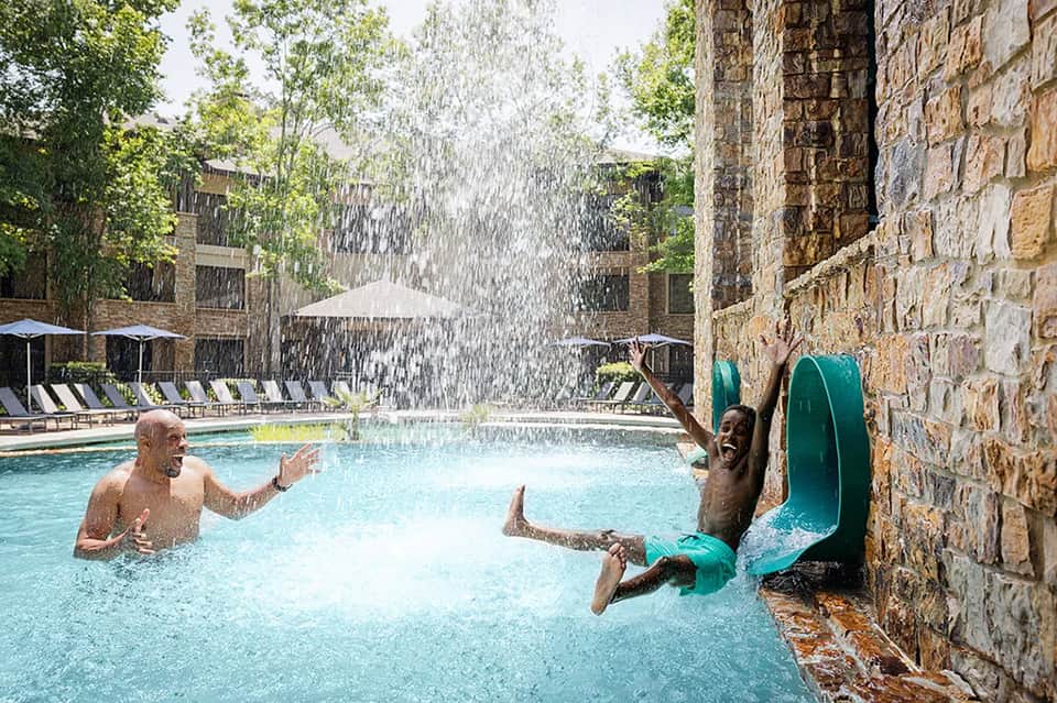 Resort pool with waterfall feature, guests swimming, stone walls, and umbrellas
