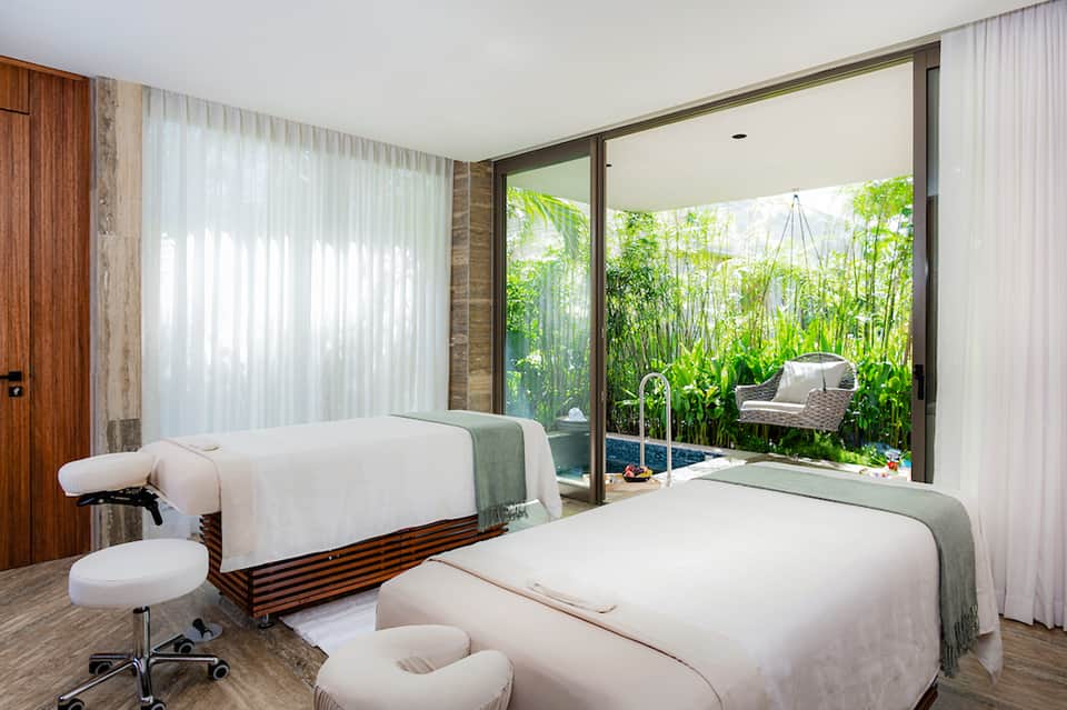 Spa treatment room with two massage beds overlooking tropical garden through large glass doors