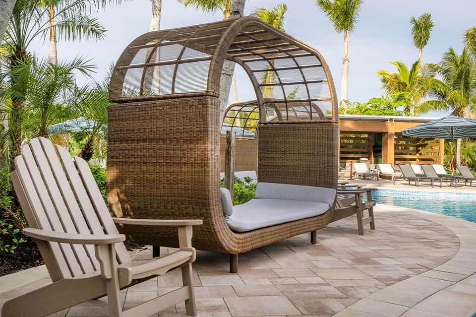Woven rattan lounge pod with gray cushion by resort pool, flanked by Adirondack chair and palm trees