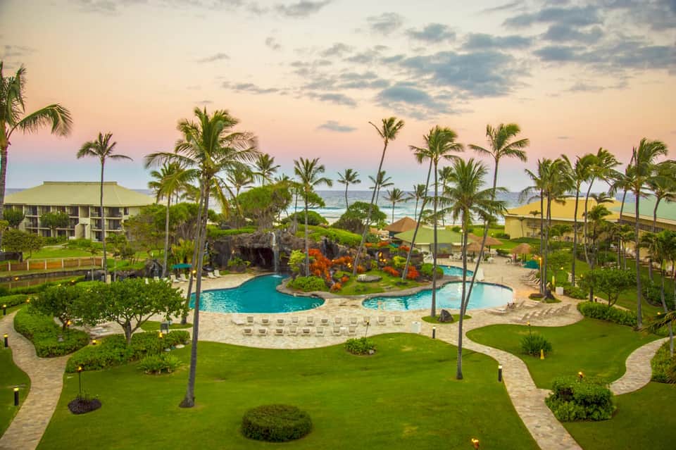 Beachfront resort grounds with curved pool, lush landscaping, palm trees, and ocean views at dusk