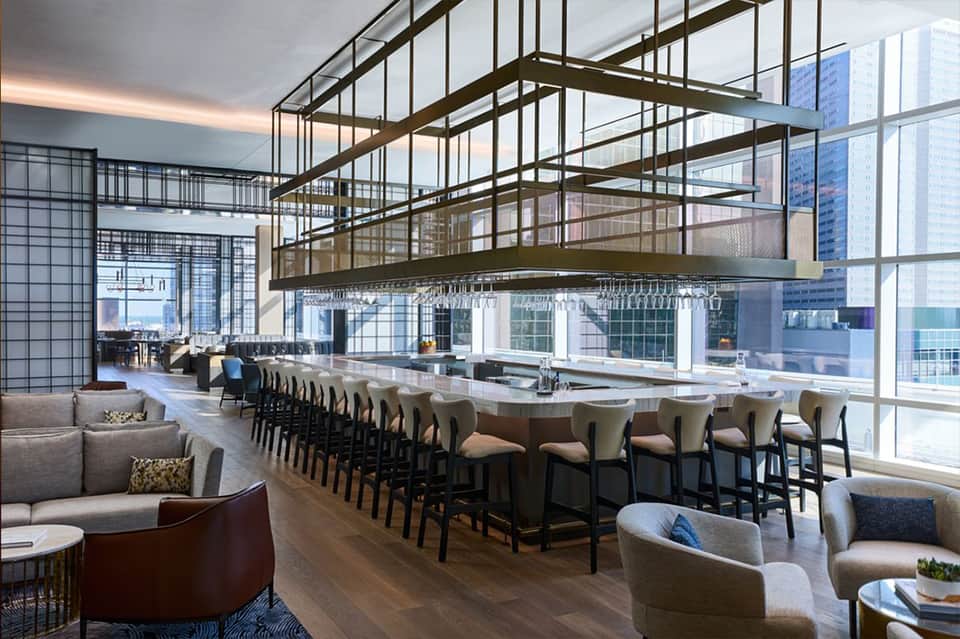 High-rise hotel bar with floor-to-ceiling windows, industrial metal ceiling framework, and city views
