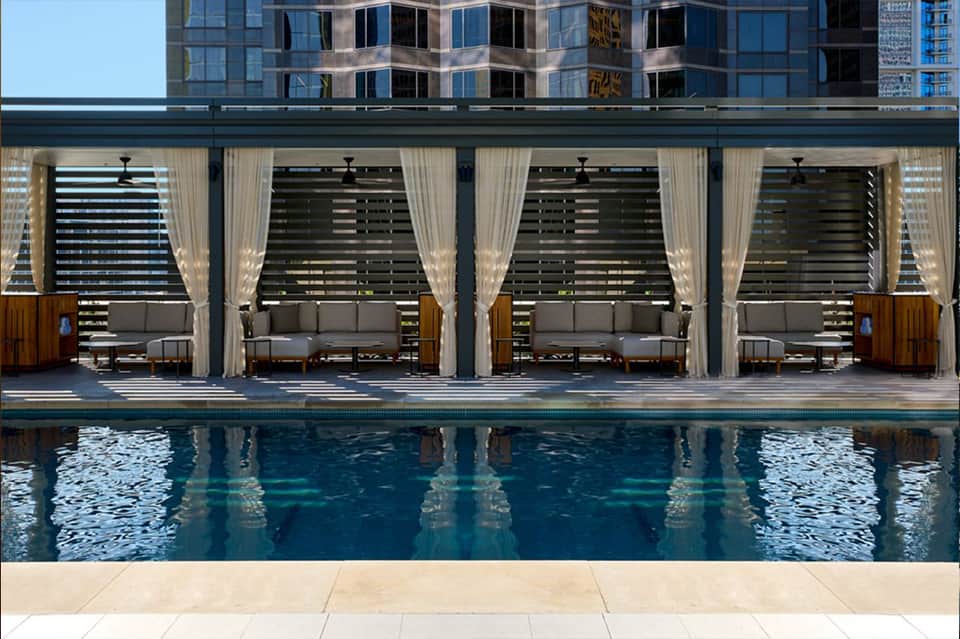 Modern indoor pool with sheer curtains, lounge cabanas, black ceiling fans, and tall glass windows