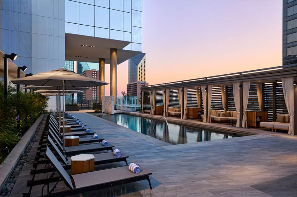 Luxury rooftop pool at sunset with lounge beds, umbrellas, cabanas, and modern city buildings surrounding