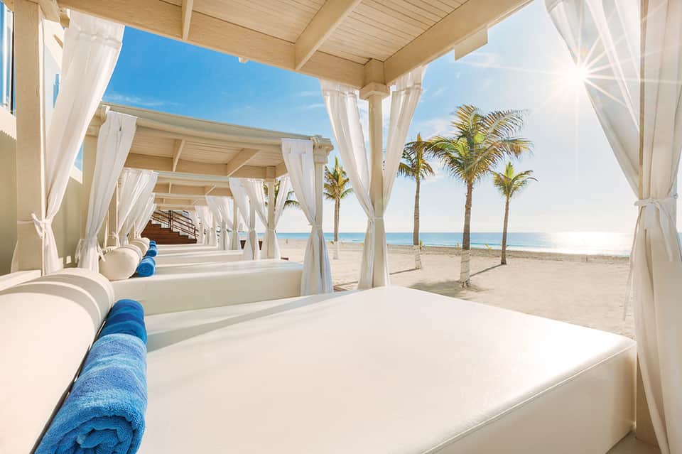 White pergola cabanas with flowing curtains along sandy beach with palm trees and ocean view
