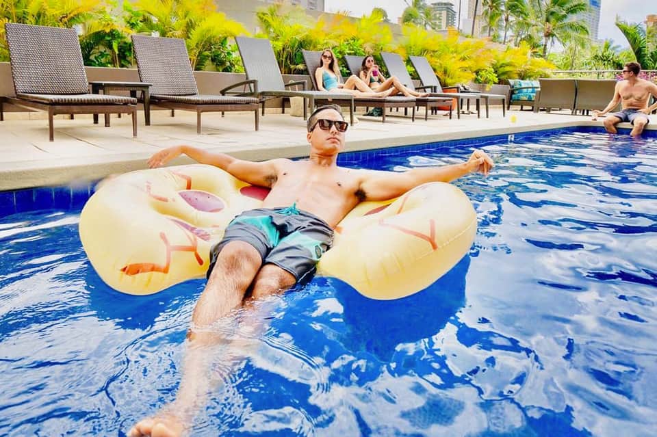 Man relaxing on yellow inflatable float in resort pool with lounge chairs and tropical plants